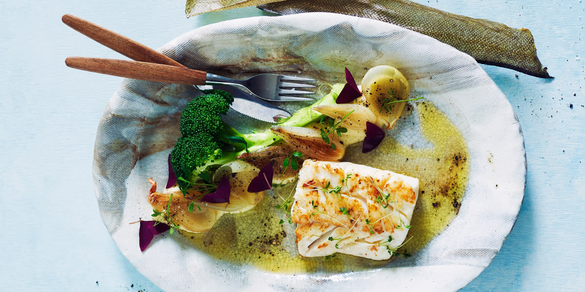 Ovnsbakt skrei med sellerirotpuré og glasert sjalottløk, servert på oval hvit tallerken med dampet brokkoli, ferske urter, kniv og gaffel.