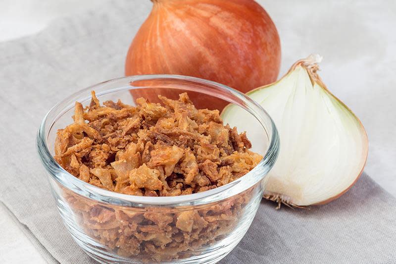 Hjemmelaget sprøstekt løk i gjennomsiktig skål av glass på beige linserviett. Hel og halvert fersk løk i bakgrunnen.