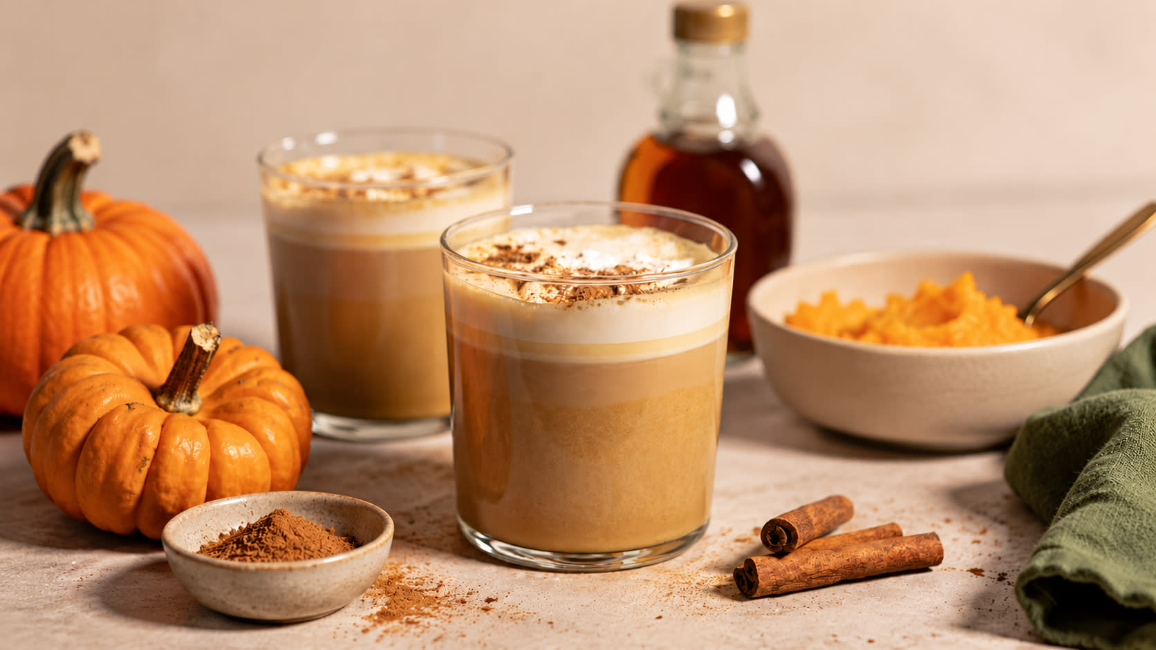 Pumpkin spice latte servert i to glass på lys kjøkkenbenk. Kanelstenger, skål med gresskarmos og liten skål med pumpkin spice rundt.