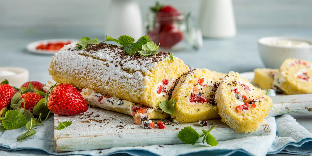 Enkel rullekake med jordbær og sjokolade, servert med krem og friske urter på lys trefjøl. Hele friske jordbær ved siden av kaken og i glass i bakgrunnen.
