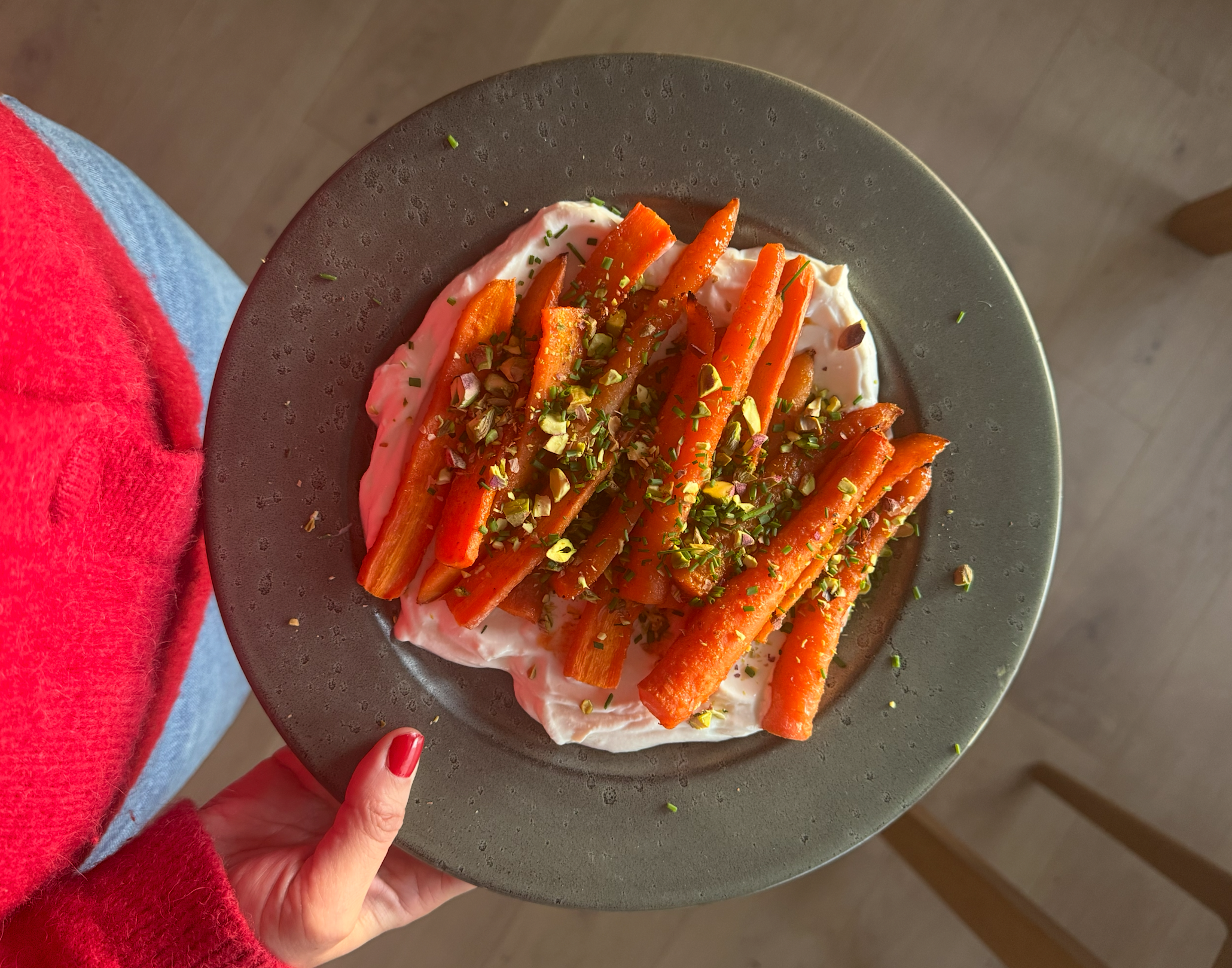 Sriracha- og honningglaserte gulrøtter servert på mørkegrå tallerken over tahini yoghurtdip, og overstrødd med hakkede pistasjnøtter. Tallerkenen holdes frem av kvinne med rød genser og rød neglelakk.