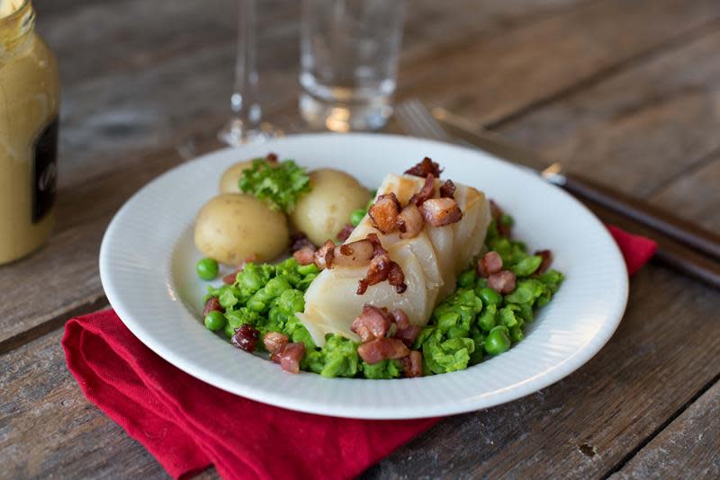 Lutefisk servert på en seng av grønn ertepuré, toppet med sprøstekt bacon. Hvit tallerken og julerød serviett på rustikk trebakgrunn.