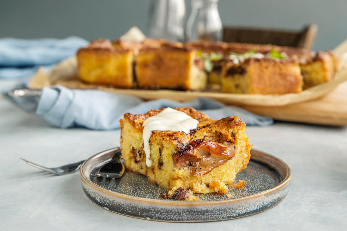 Klassisk eplekake i langpanne servert i stykke med vaniljesaus på toppen, på grå tallerken på dekket bord. Oppdelt eplekake i langpanne i bakgrunnen.