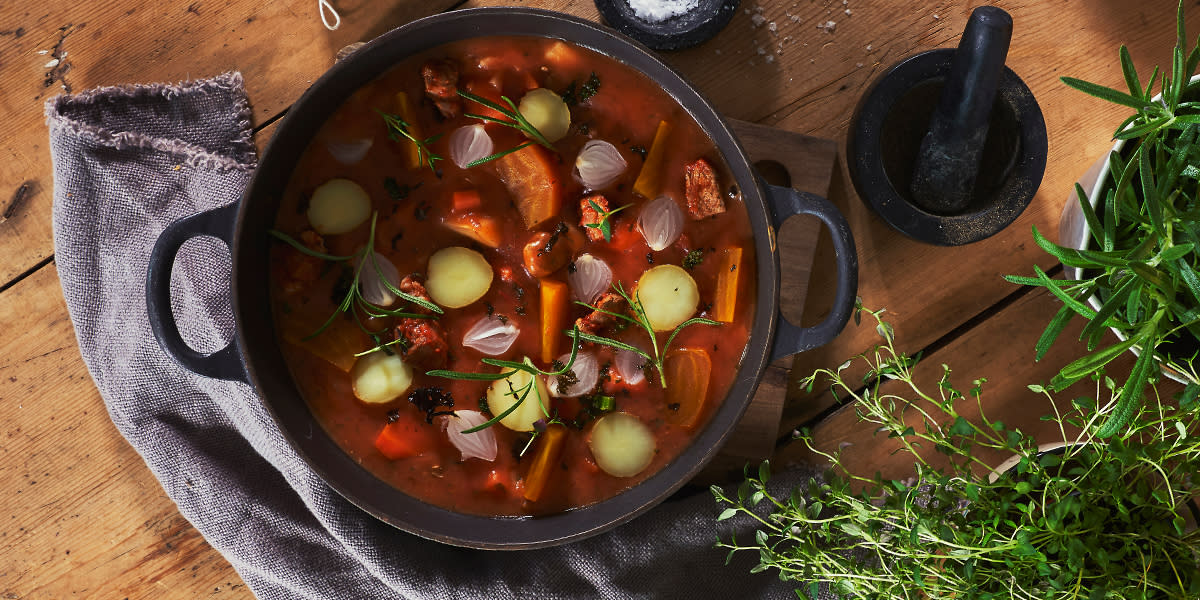 Provencalsk lammegryte med lammekjøtt, poteter, løk, gulrøtter og tomater servert i svart støpejernsgryte på rustikk trebord. Ferske urter ved siden av.