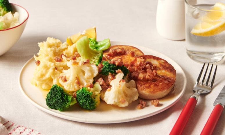 Fiskekaker med potetmos og bacon, servert med blomkål, brokkoli og brunet smør på hvit middagstallerne på beige dekket middagsbord. Rød kniv og gaffel ved siden av.