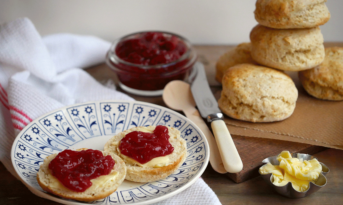 Perfekte luftige scones med jordbærsyltetøy servert på blåmønstret hvit asjett på mørkt trebord. Stabel med scones, og glass med jordbærsyltetøy i bakgrunnen.