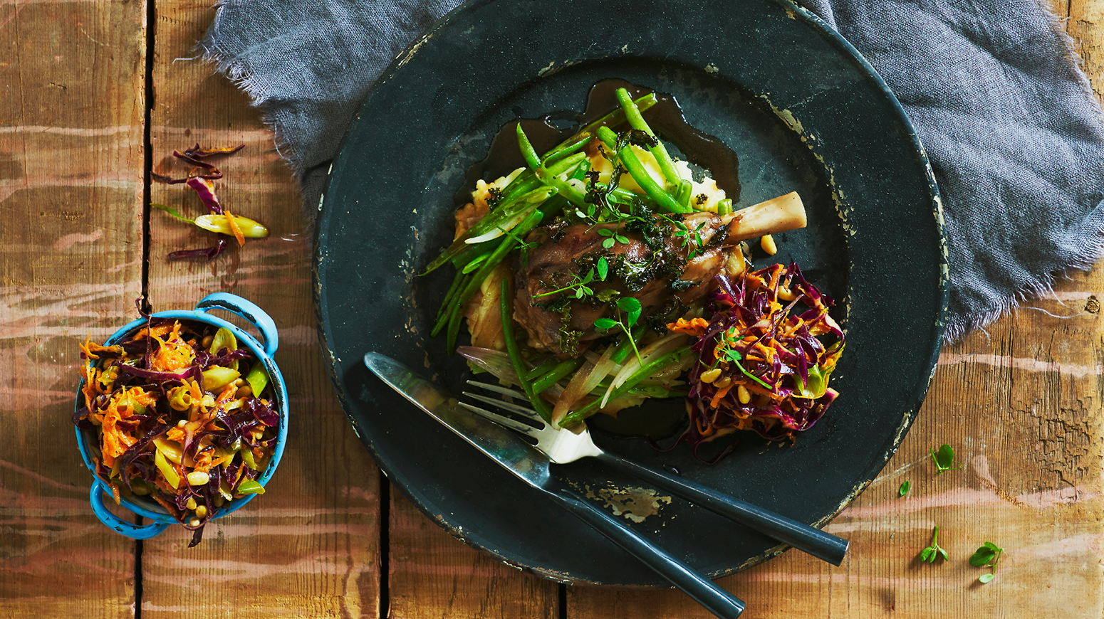 Rødvinsbrassert lammeskank med potetpuré, aspargesbønner, rødkålsalat og dressing servert på mørk tallerken, på rustikk trebord. Grå linserviett under tallerkenen.