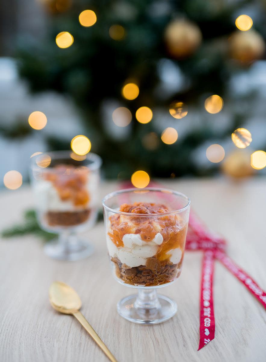 Ostekake på glass med pepperkaker og multer servert på trebord med gullskje ved siden av. Juletre med lys i bakgrunnen.