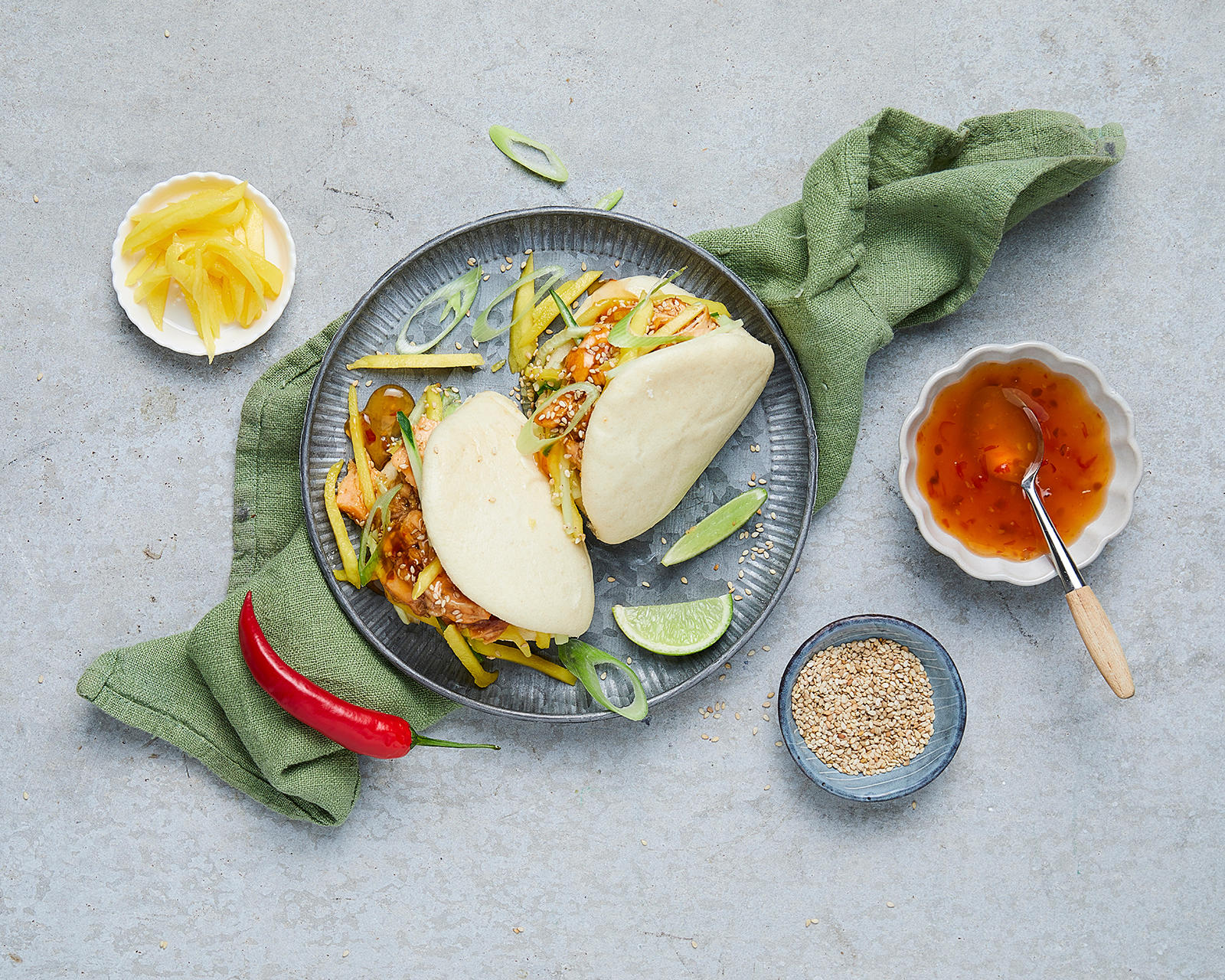 2 stk steam buns med teriyakilaks og mango på en blå tallerken med mango, sesamfrø og chillisaus i skåler rundt.