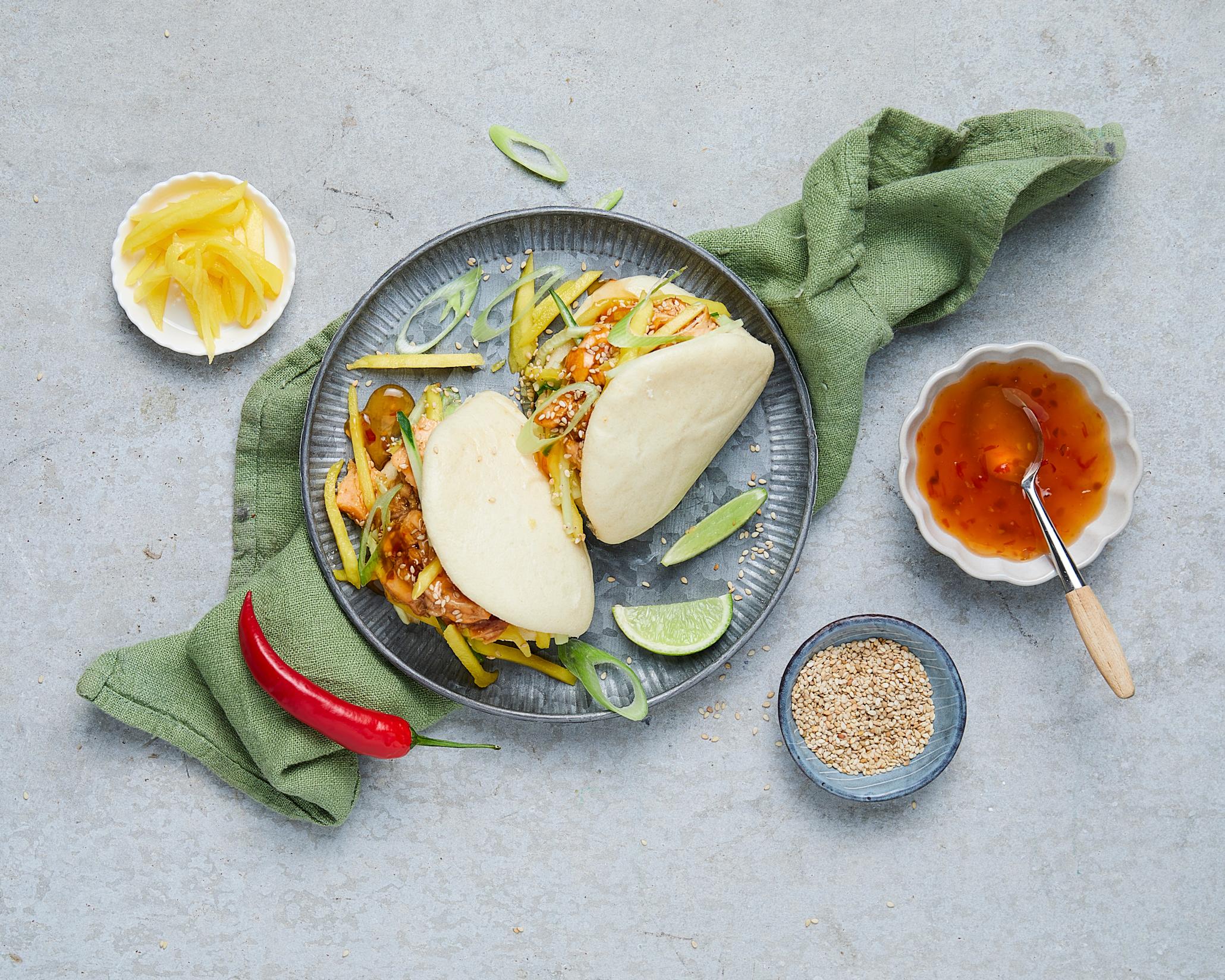 2 stk steam buns med teriyakilaks og mango  på en blå tallerken med mango, sesamfrø og chillisaus i skåler rundt. 
