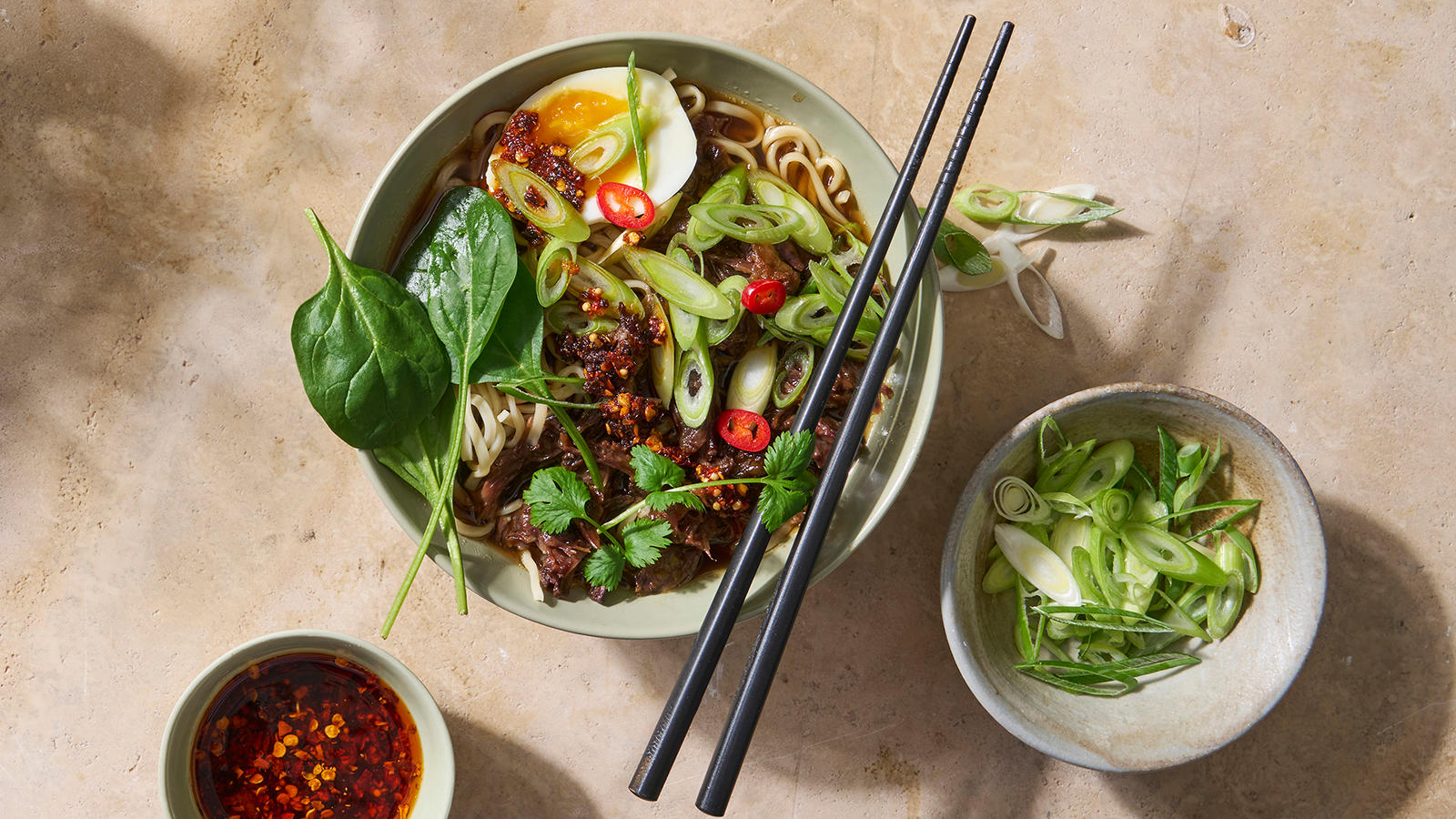 Ramen med oksehale og chilicrisp. En keramisk bolle med ramen fylt med mørt oksehalekjøtt, nudler og fyldig kraft, toppet med egg, vårløk, spinat, koriander, rød chili og chilicrisp. Ved siden av står det to mindre skåler med vårløk og chilicrisp.