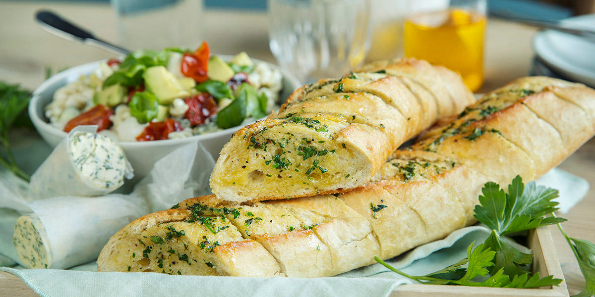 Hjemmelaget hvitløksbaguette servert på trebrett med hjemmelaget hvitløkssmør, og en frisk salat i bakgrunnen.