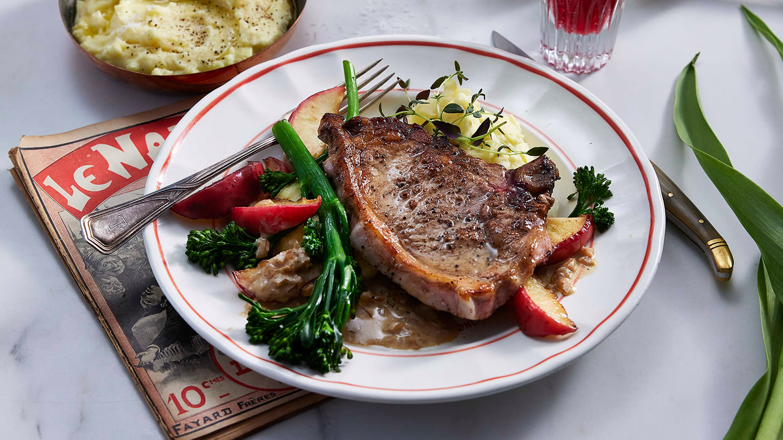 Svinekoteletter med kremet eplesaus og potetstappe servert på hvit tallerken med rød kant, med brokkolini og friske urter.