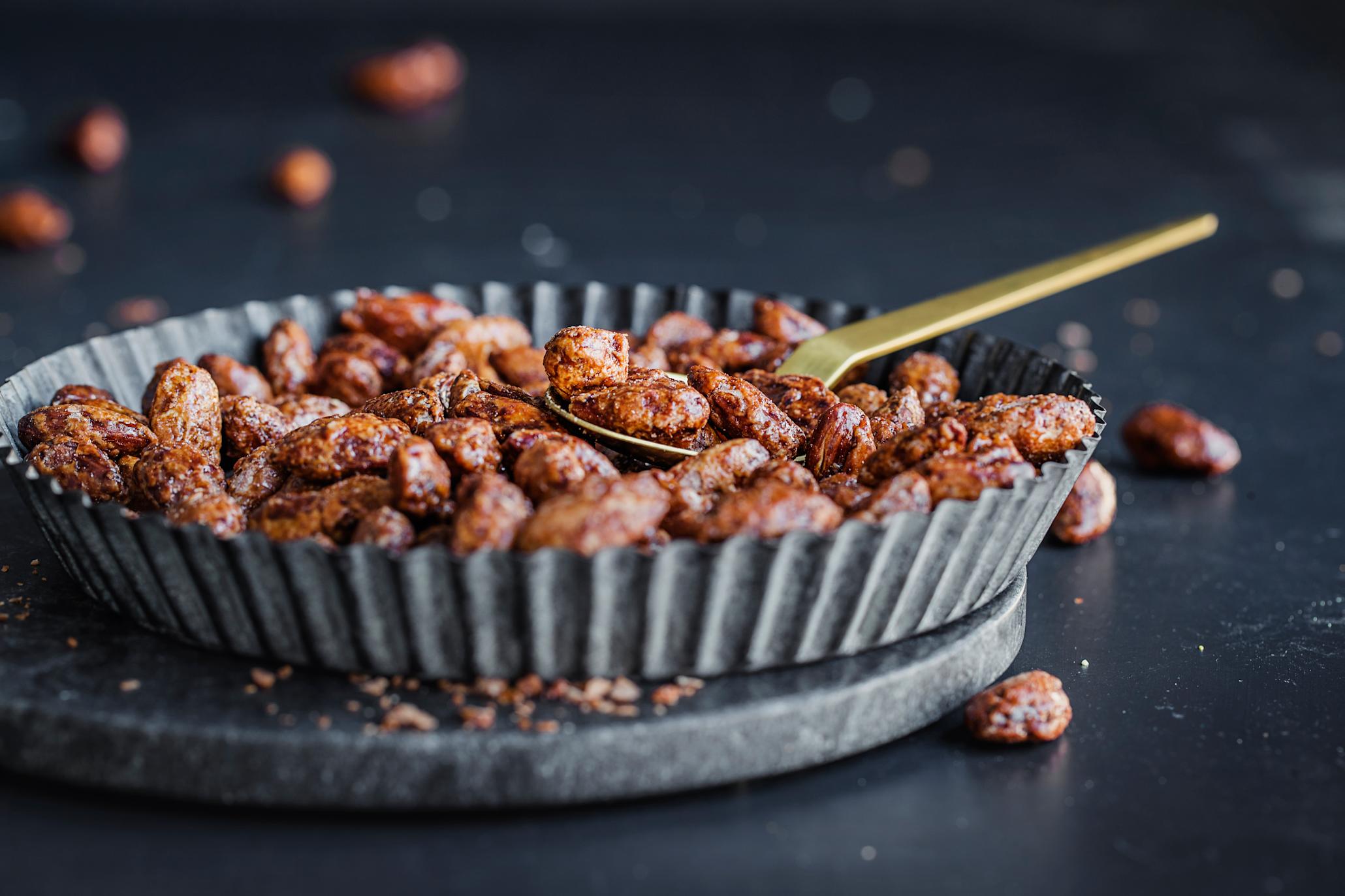 Brente mandler med kanel servert i grå metallform med riller, med gullskje stukket inn blant mandlene.