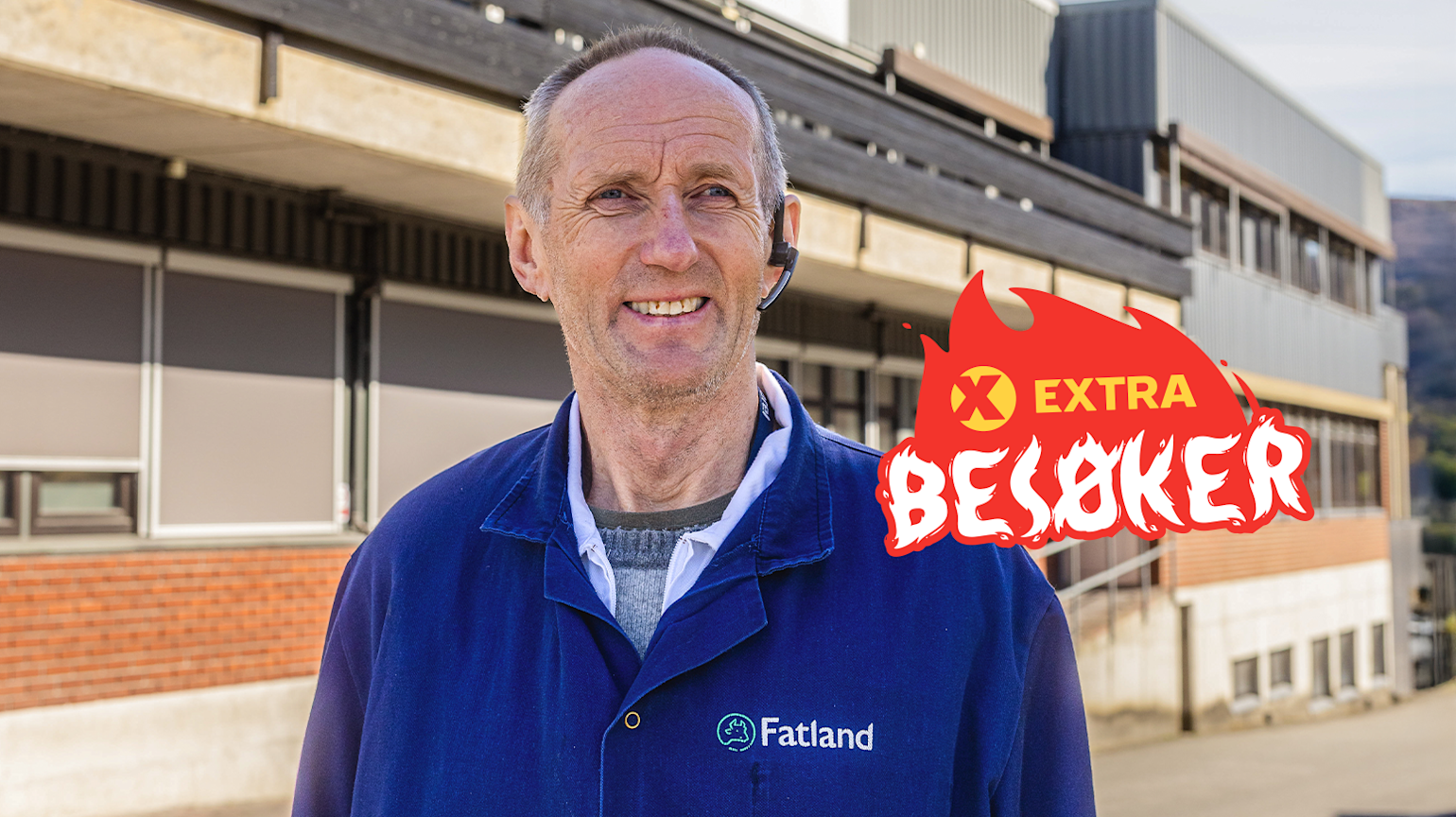 En mann med blå jakke med "Flatland" logo på står foran en fabrikk. "Extra Besøker" log er på bilde.