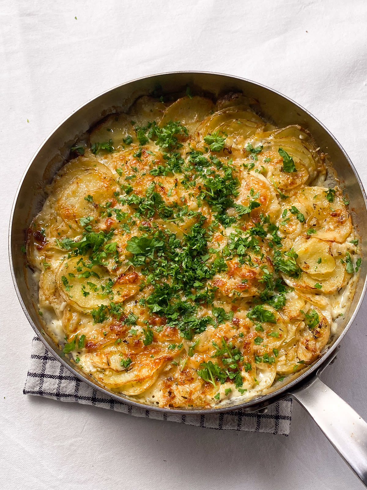 Fløtegratinerte poteter med parmesan pyntet med grønne urter og servert i stekepanne i metall på rutete linserviett.