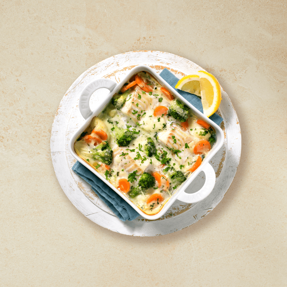 Ovnsbakt torsk i hvit saus i en hvit ildfast form med brokkoli og gulrot, servert med sitronbåter på hvit asjett. Beige bakgrunn.
