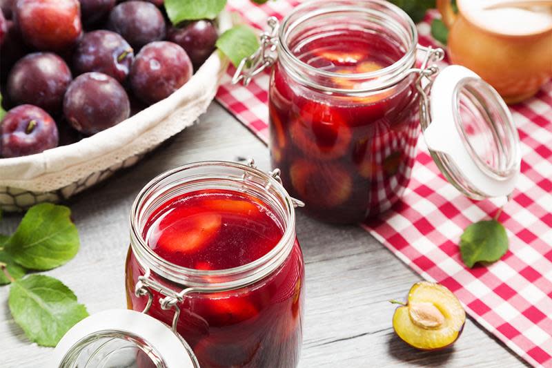 To store Norgesglass med hermetiserte plommer, på rustikk trebord med rødrutet duk. Kurv med plommer i bakgrunnen.