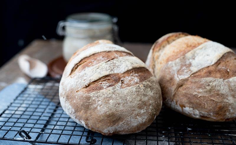 Hjemmelaget surdeigsbrød servert på rist, på trebord. Brødene har mørk skorpe, og en melet overflate. Glass med surdeigstarter i bakgrunnen.