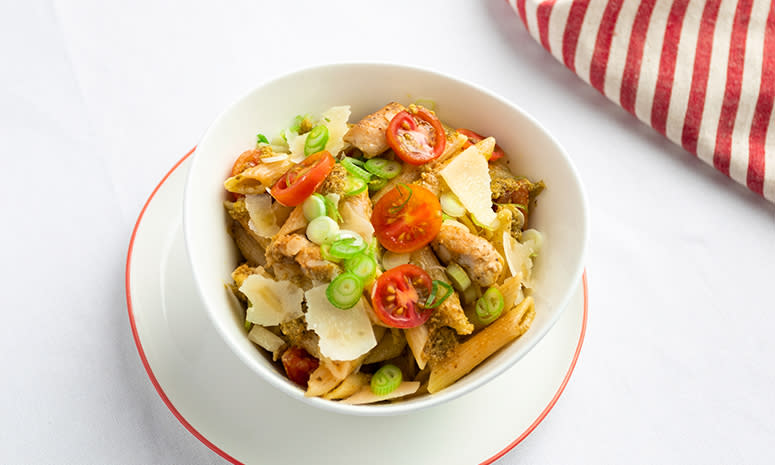 Pasta med kylling, pesto og oppskjært tomat, servert i en hvit skål som står på en hvit tallerken med rød kant, ligger på en hvit duk