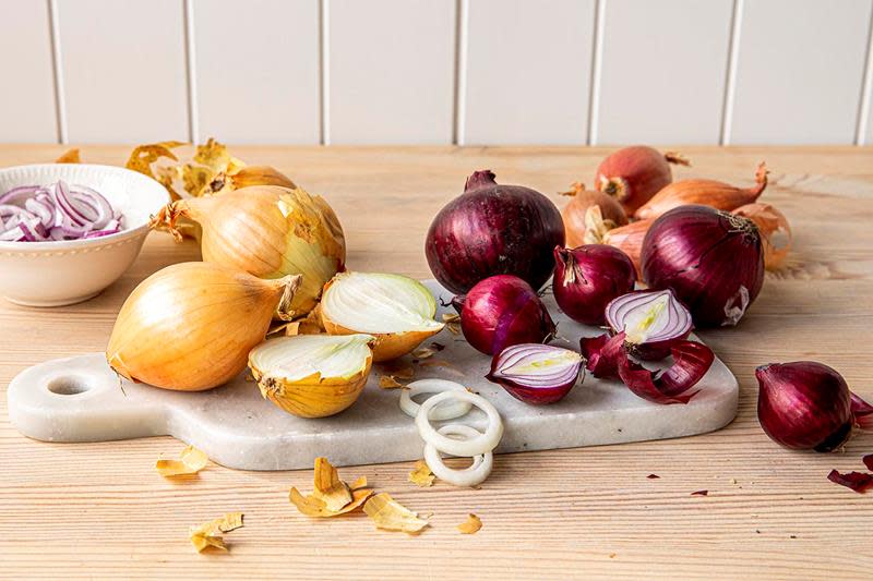 Løk i forskjellige sorter og farger ligger på kjøkkenbenk med fjøl i marmor. Rødløk, guk løk og sjalottløk, både hel, i båter og i ringer.
