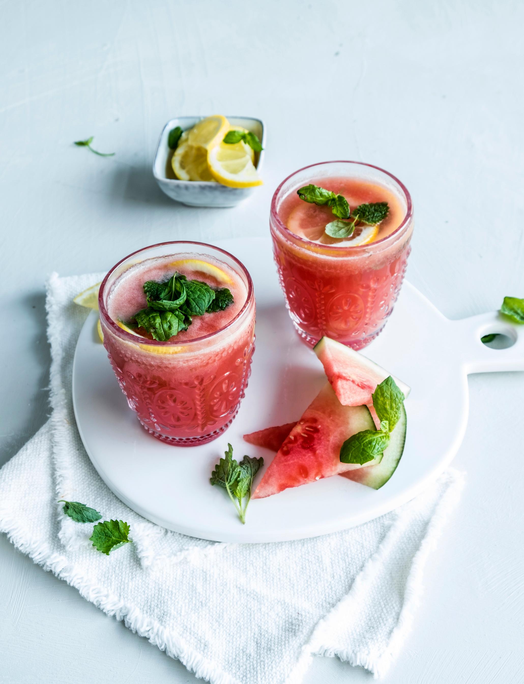Vannmelon- og ananascooler pyntet med frisk mynte og servert i mønstrete glass på hvit marmorfjøl med vannmelonskiver ved siden av. Liten skål sitronskiver i bakgrunnen.