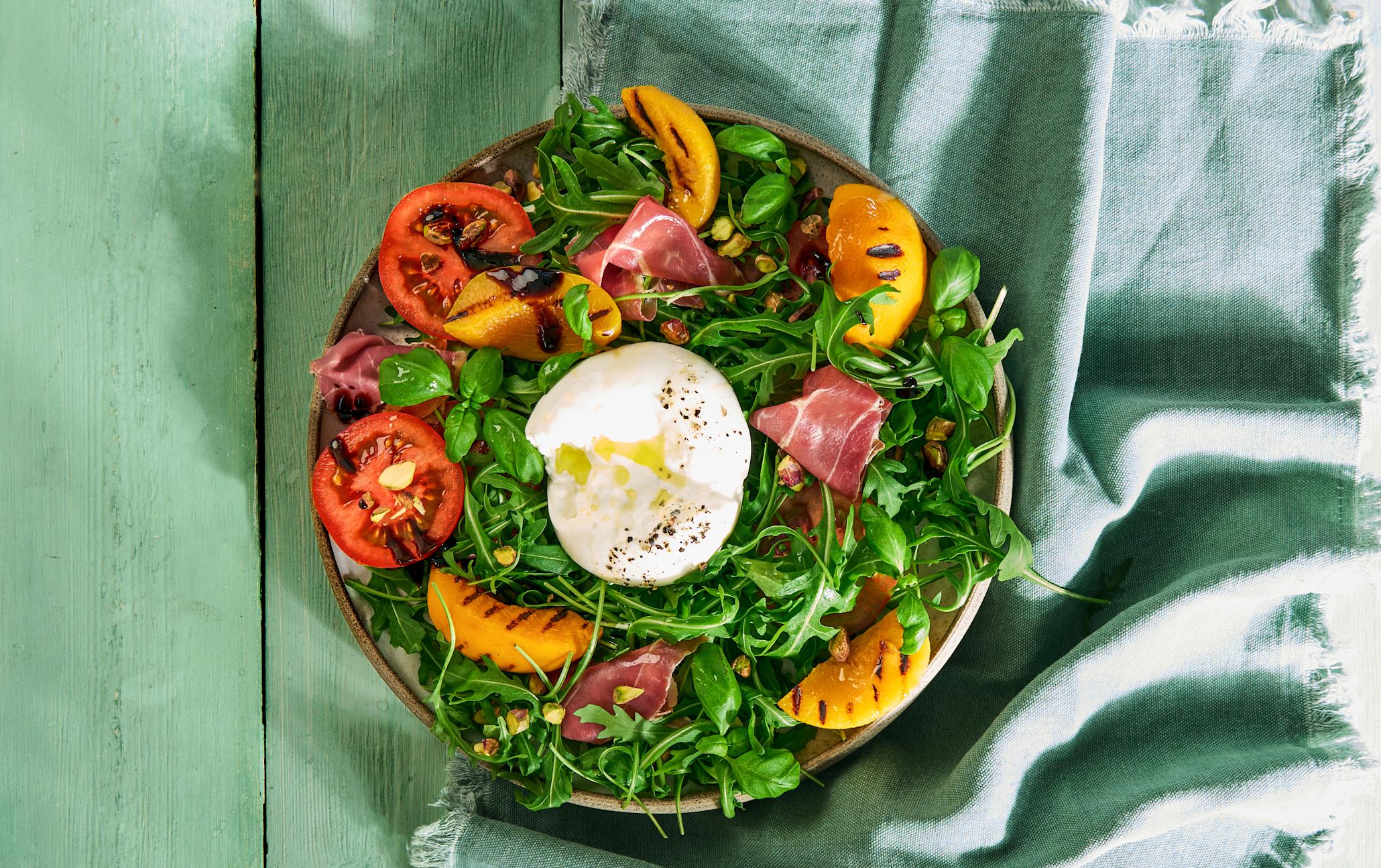 Sommersalat med grillet fersken, spekeskinke og burrata, servert i skål med ruccola, tomater og pistasjenøtter, på grønt trebord.