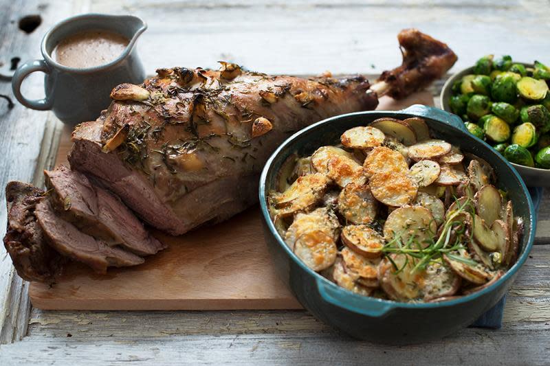 Langtidsstekt lammelår spekket med hvitløksfedd, servert på trefjøl skåler med gylne fløtegratinerte poteter og rosenkål. Liten mugge med saus på siden.