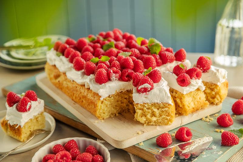 Saftig langpannekake med kokos servert på trefjøl, pyntet med sveitsisk marengskrem, ferske bringebær og sitronmelisse. Kaken står på dekket bord, med asjetter og en vannkaraffel i bakgrunnen.