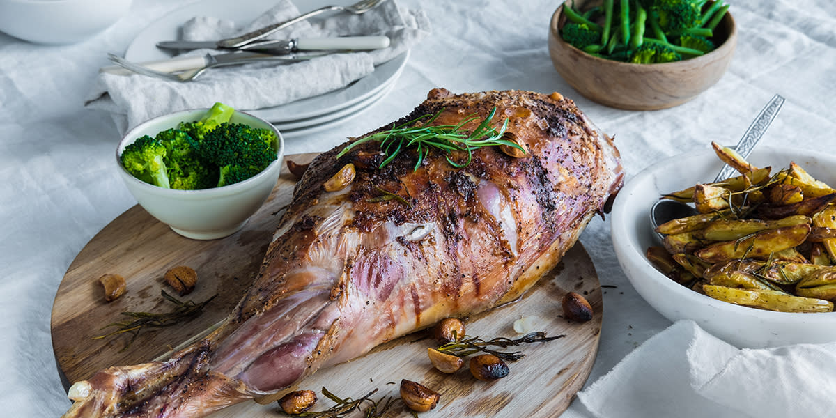 Helstekt lammelår servert på rund trefjøl, på hvit duk. Hvit skål med ovnsstekte potetbåter, liten hvit skål med brokkoli, og medium stor treskål med grønne bønner ved siden av.