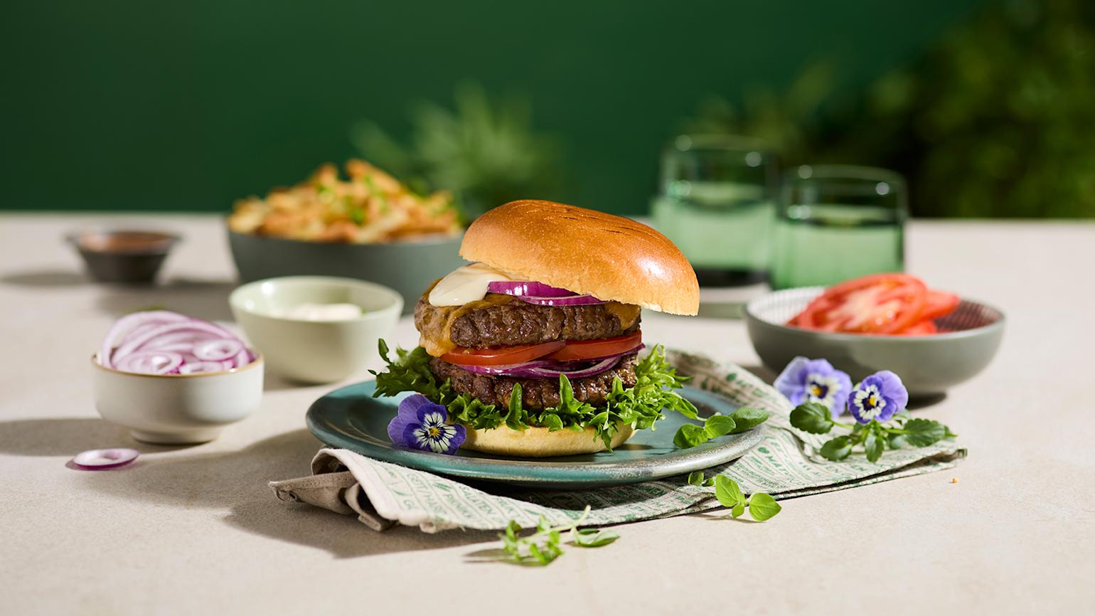 Coop Mega - Dobbel steakburger med cheddar og aioli servert på sjøgrønn asjett på beige bord. Skåler med tilbehør rundt.