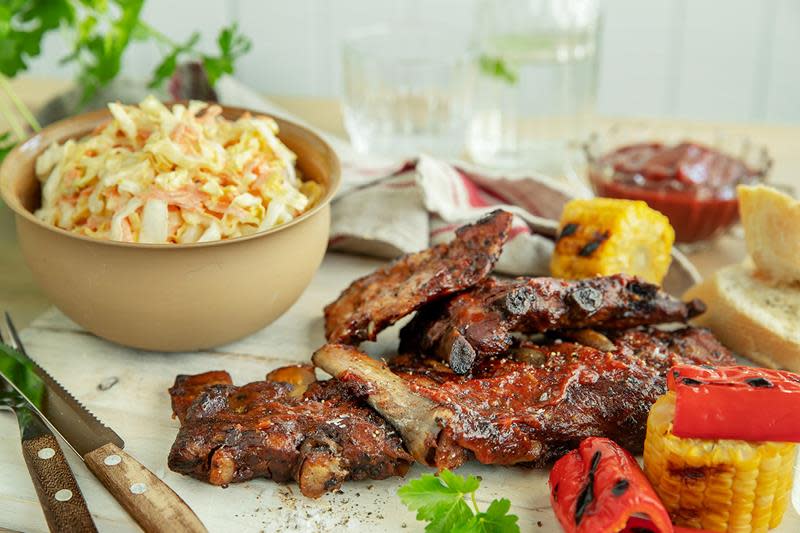 Nygrillede spareribs servert på hvit trefjøl, med grillet mais, paprika og ferske urter. Hjemmelaget barbecuesaus og hjemmelaget coleslaw ved siden av.