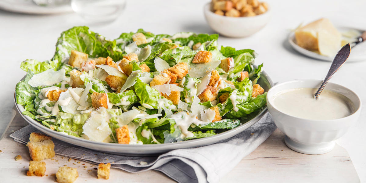 Cæsarsalat med hjemmelagde krutonger, parmesan og dressing, servert på en stor lys tallerken. Skål med dressing ved siden av.