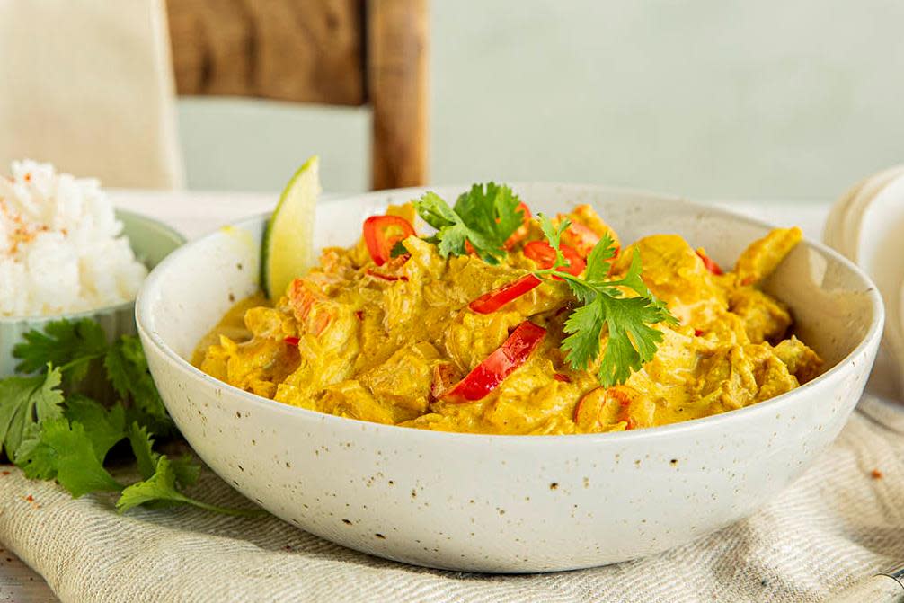 Knall gul kremet karrigryte med rød paprika og rester av kalkun servert i lys skål. Limebåter, fersk koriander og ris på siden. Lys bakgrunn.