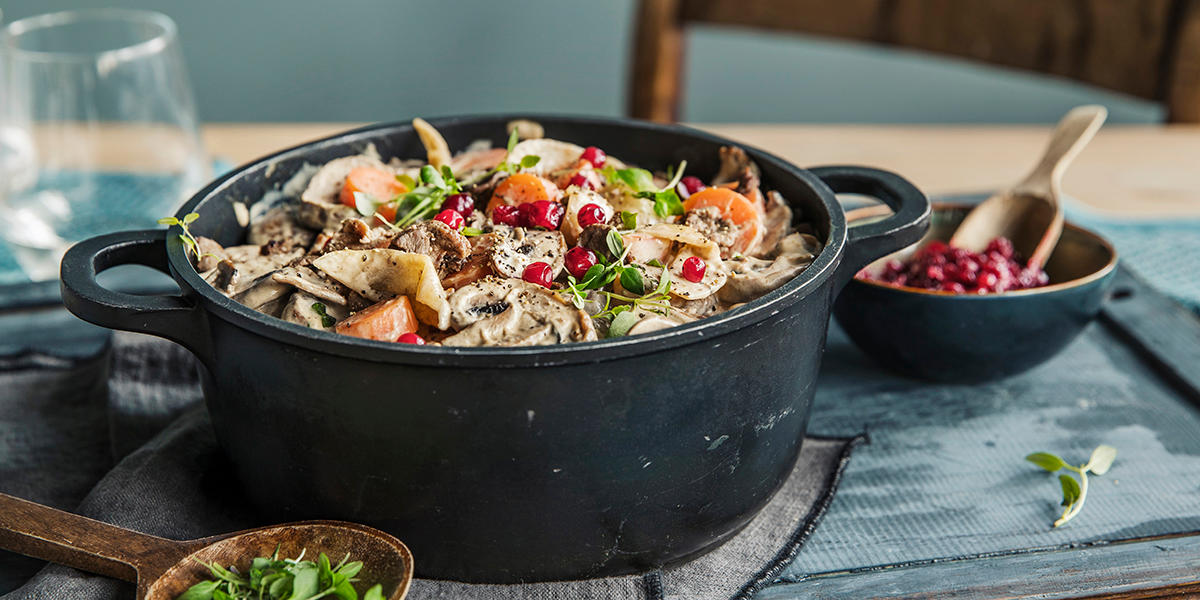 Finnbiff med sopp, gulrot og løk i rustikke biter, servert i svart støpejernsgryte med tyttebær og ferske urter på toppen. Gryten står på et rustikk dekket middagsbord, med en liten skål tyttebærsyltetøy ved siden av, og en mørk tresleiv med ferske urter.