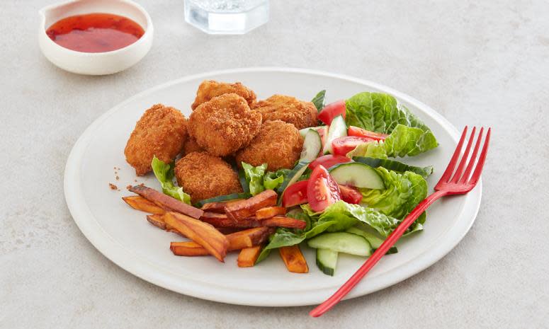 Crispy fiskenuggets med ovnsbakt søtpotet, på hvit tallerken med salat, tomat og agurk. Rød gaffel på siden. Lys bakgrunn.