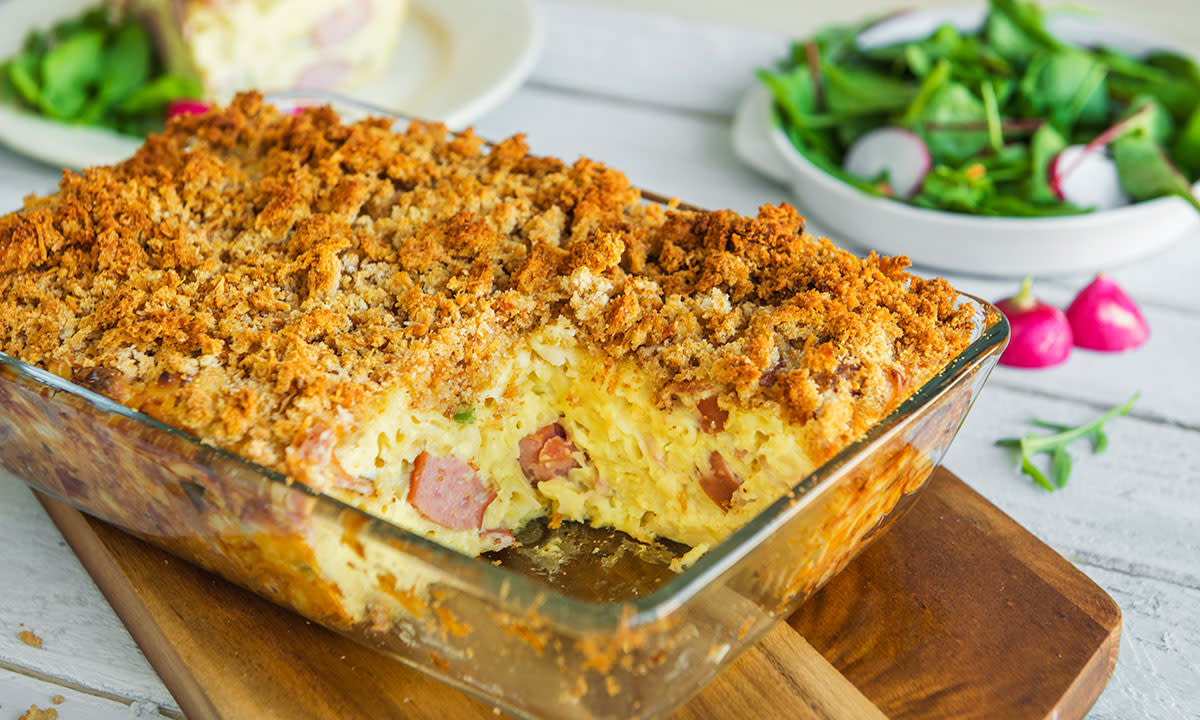 Makaronigrateng med pølse og sprø topping, bacon og cheddar servert i gjennomsiktig ildfast form på trefjøl på middagsbord. Frisk grønn salat med redikker i bakgrunnen.