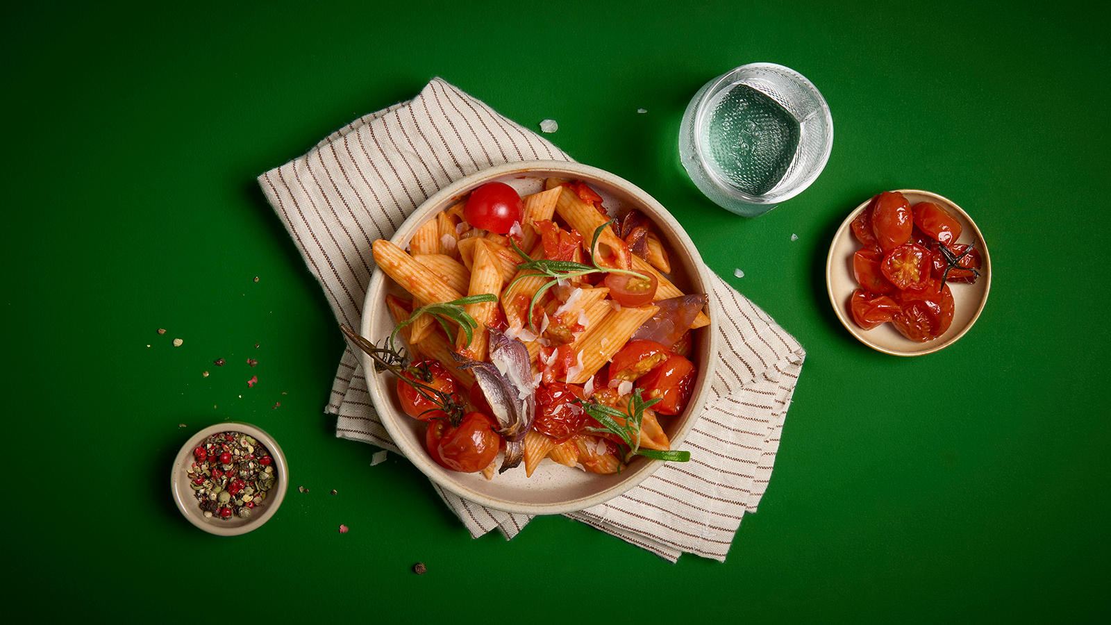 Pasta penne med ovnsbakte tomater servert i liten skål med frisk rosmarin på toppen. Små skåler krydder og ovnsbakte tomater ved siden av.