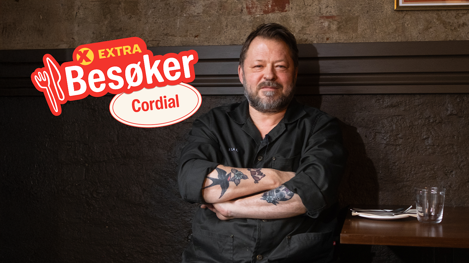 Mann i svart kokkejakke med tatoveringer står med armene i kors. "Extra Besøker" Cordial-logo på bilde.