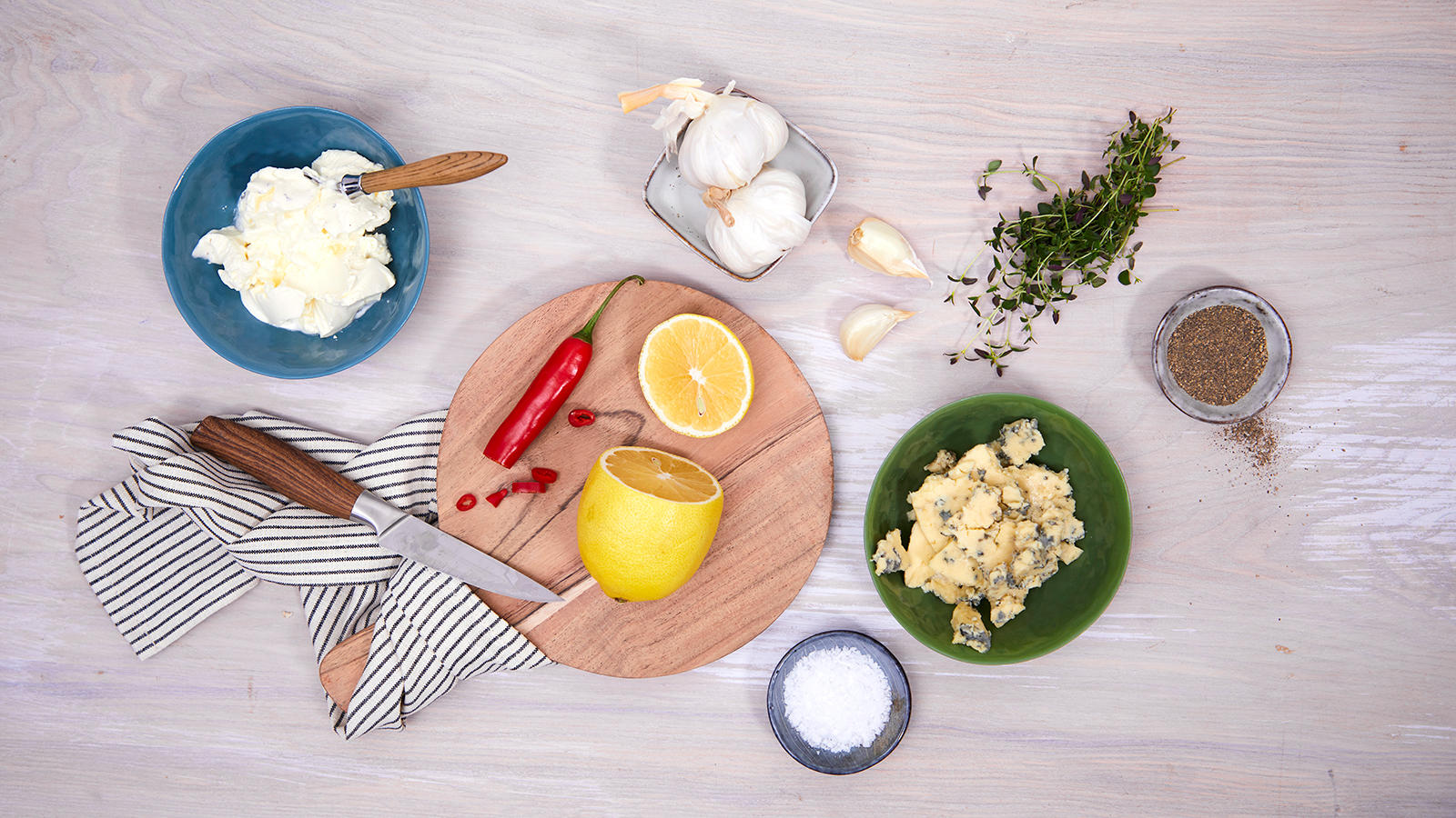 Hvit saus til pizza illustrert med ingredienser som creme fraiche, sitron, chili, hvitløk og blåmuggost i skåler og på fat på et lyst trebord.