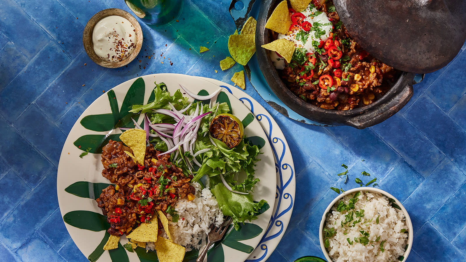 Chili con carne - meksikansk gryte, servert på mønstret tallerken med crispisalat, rødløk, ris og tortillachips. Gryte med chili con carne ved siden av.