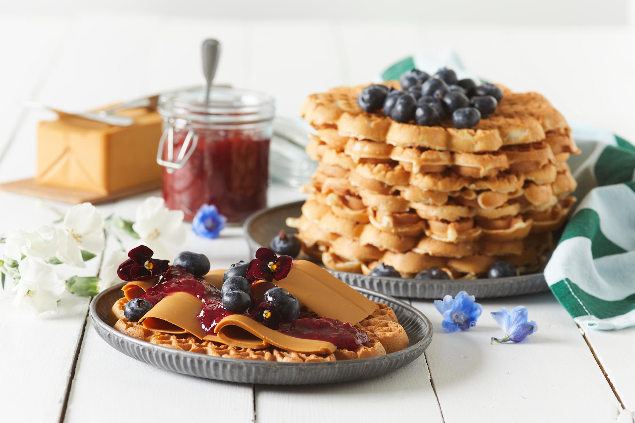 Vafler uten egg servert på mørkegrå tallerken med brunost, syltetøy, blåbær og blomster. Brunost, glass med syltetøy, og mørkegrå tallerken med en stabell vaffel i bakgrunnen.