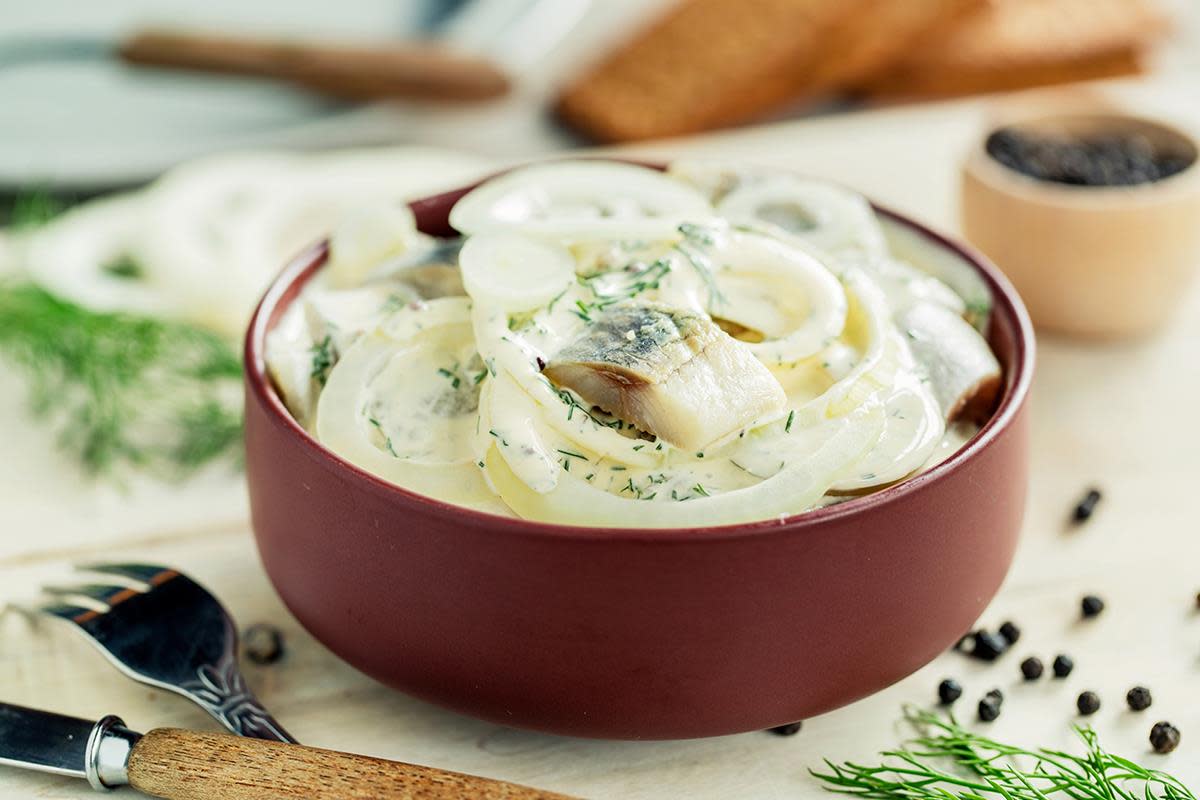 Hjemmelaget rømmesild med løk og dill, servert i brunrød skål på lys trebakgrunn. Fersk dill og hele pepperkorn strødd rundt.