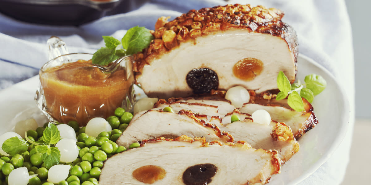 Fylt svinestek med sprø svor, eplesaus og karamelliserte poteter servert på hvitt ovalt fat med sølvløk, erter og ferske urter.