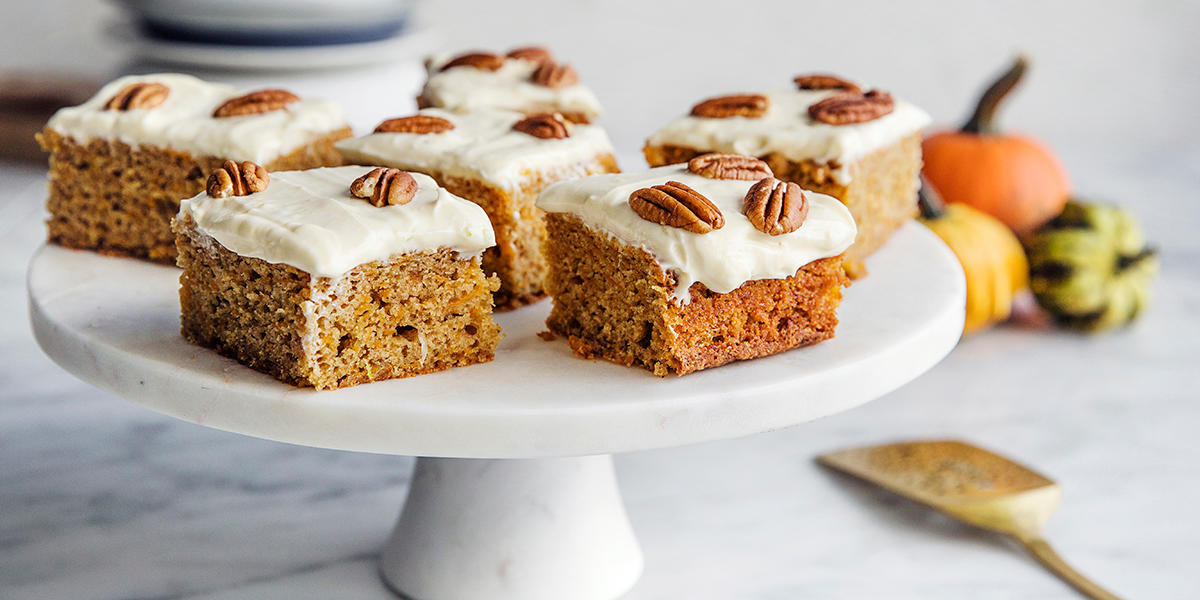 Gresskarkake med ostekrem delt i biter, pyntet med pekannøtter og servert på hvitt kakefat i marmor. Små pyntegresskar i bakgrunnen.