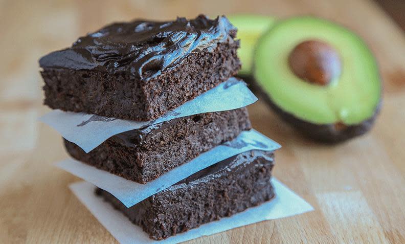 Fudgebrownies med avokado pyntet med sjokoladeglasur, delt opp i ruter, og stablet med matpapir mellom. Lyst treunderlag, med en halv avokado i bakgrunnen.