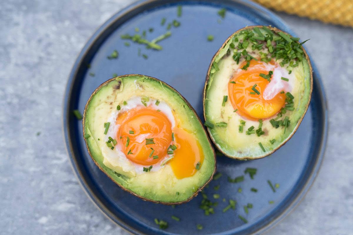 Bakt avokado delt i to, med egg i midten og ferske urter drysset over, servert på blå tallerken.