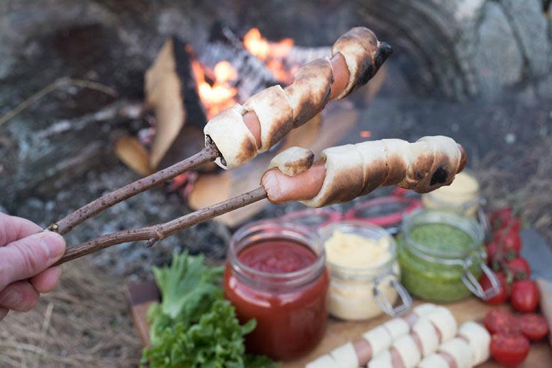 Innbakte pølser på tur, grillet på bål. I bakgrunnen synes forskjellige krukker med dipsaus, og noen grønnsaker på trefjøl, samt flere pølser med deig surret rundt som ikke er grillet enda.