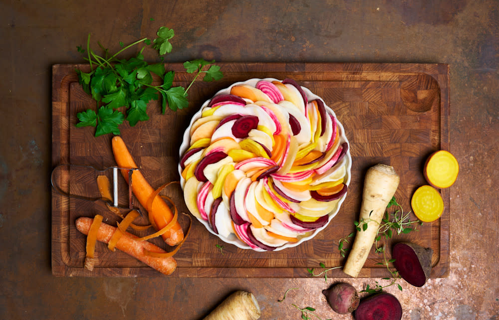 Ovnsbakte rotgrønnsaker med urtesmør og parmesan servert i en hvit paiform på en skjærefjøl av mørkt tre. Rotgrønnsaker og friske urter rundt.
