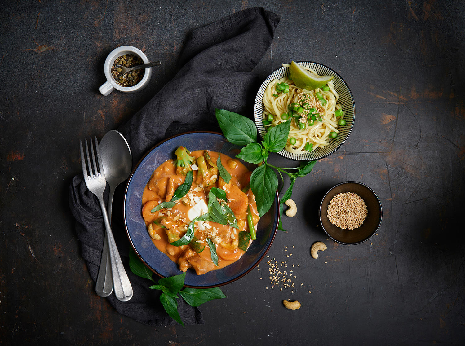 Rød curry med svinekjøtt og thai basilikum servert i mørk skål med frisk thai basilikum rundt. To mindre skåler med sesamfrø og nudler ved siden av.